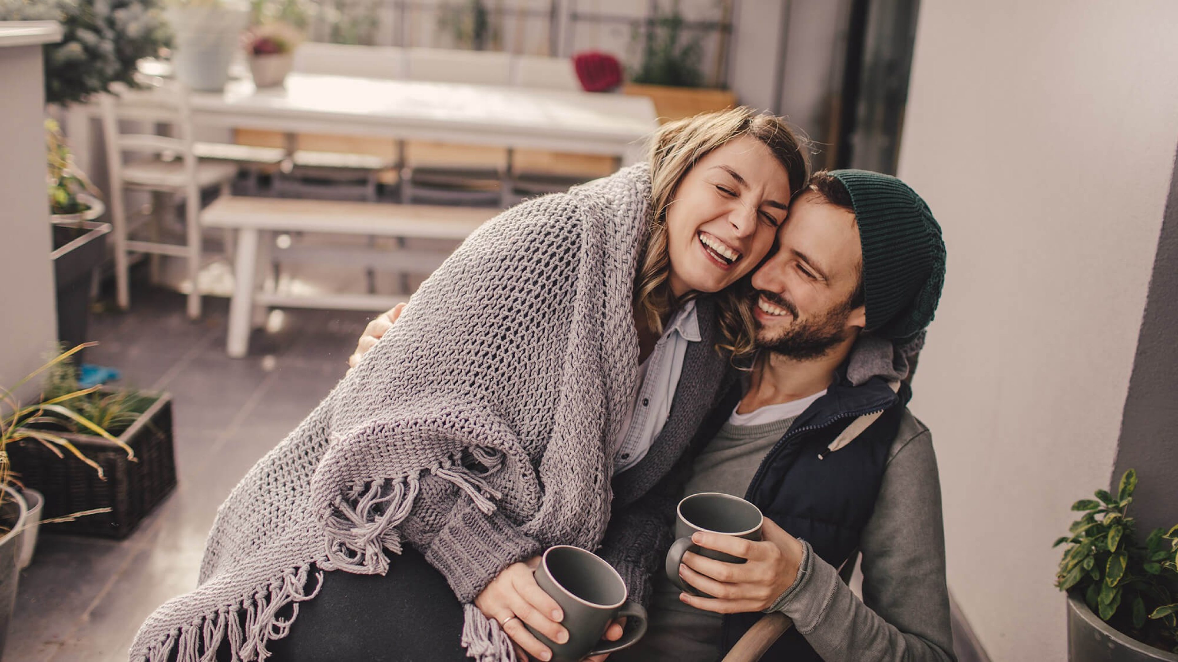 Junges Paar lachend auf einem Balkon, sie sitzt auf seinem Schoß, beide sind warm angezogen und haben eine Kaffeetasse in der Hand