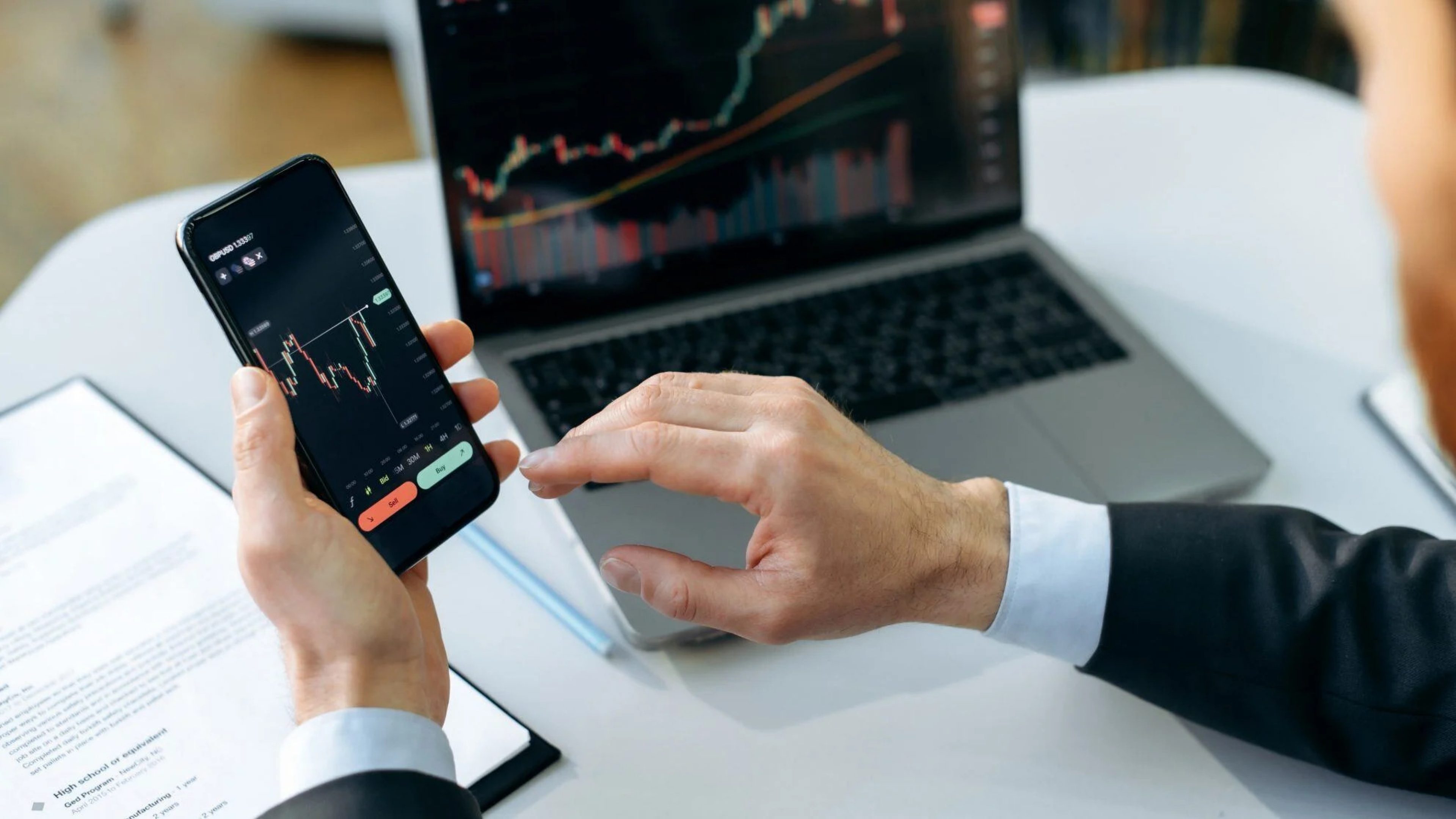 Geschäftsperson analysiert Finanzdiagramme auf Smartphone und Laptop am Arbeitsplatz.