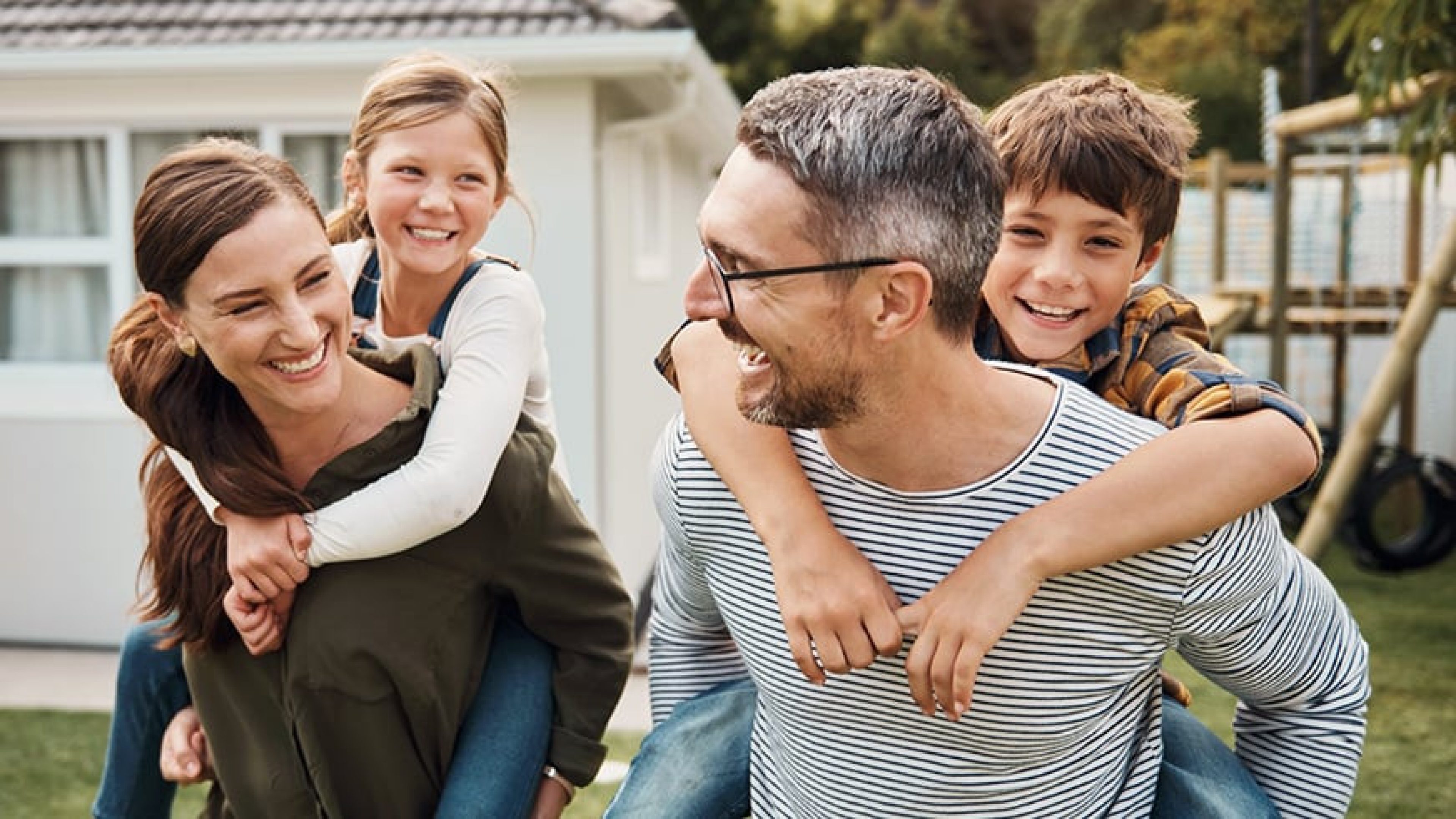 Glückliche Familie mit zwei Kindern beim Huckepack-Spiel im Garten vor ihrem Zuhause.