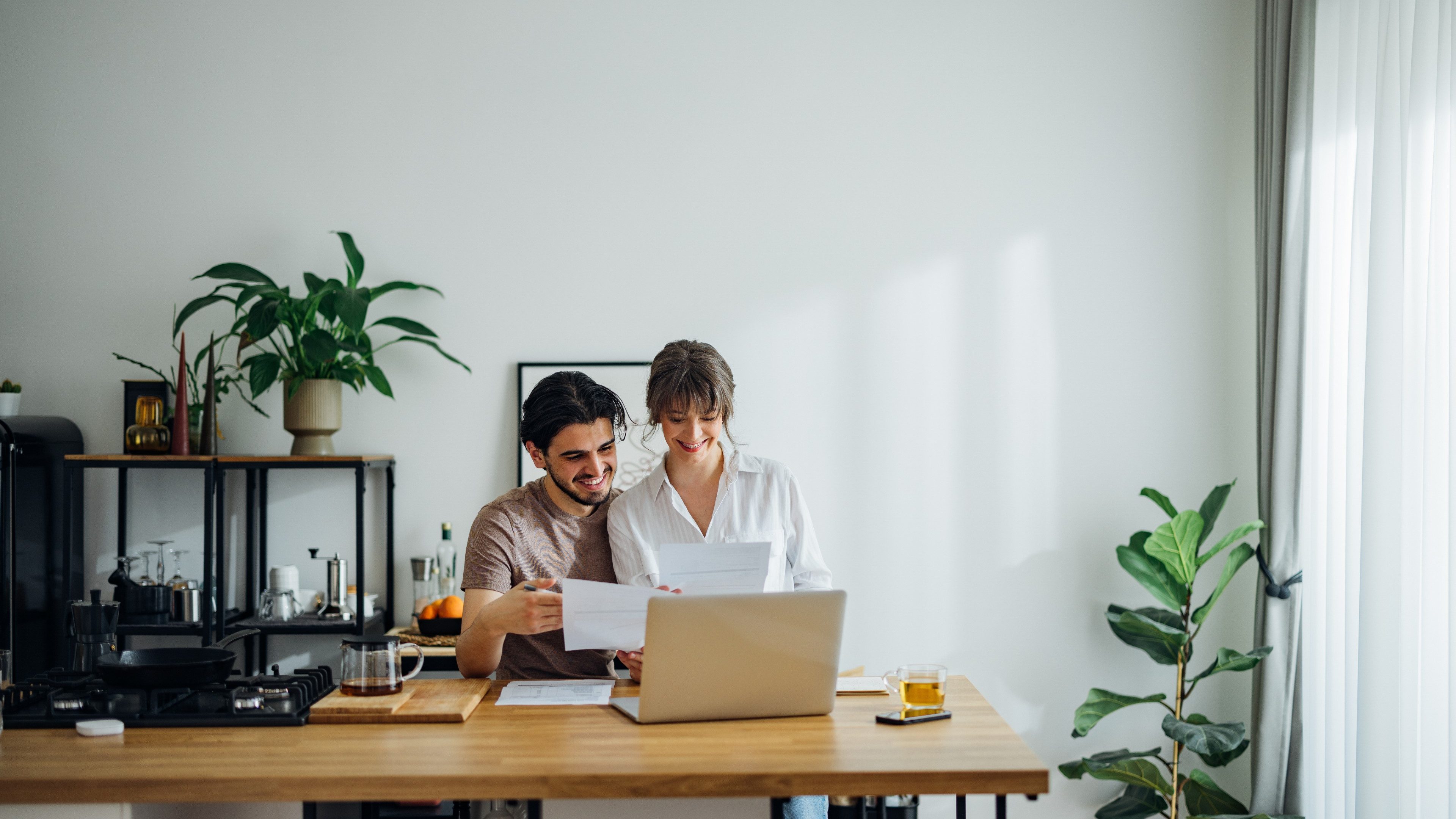 Happy boyfriend and girlfriend laughing while doing home finances together online on a laptop computer in the kitchen.