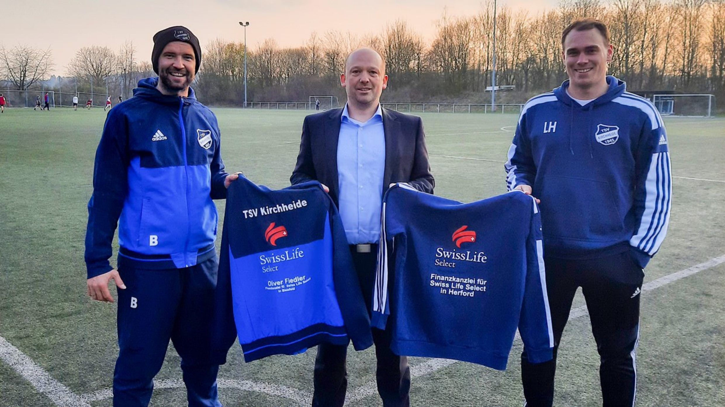 Finanzberater Oliver Fiedler  zeigt mit einem Spieler und Trainer ein Fußballtrikot mit Swiss Life Select Logo